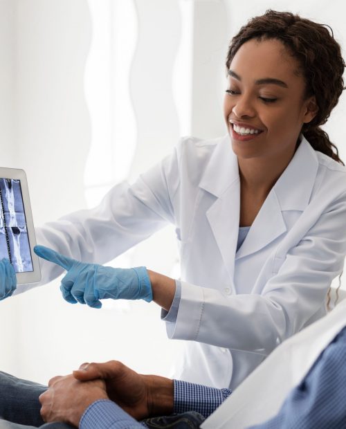Smiling african female dentist showing young man patient teeth xray, using digital tablet